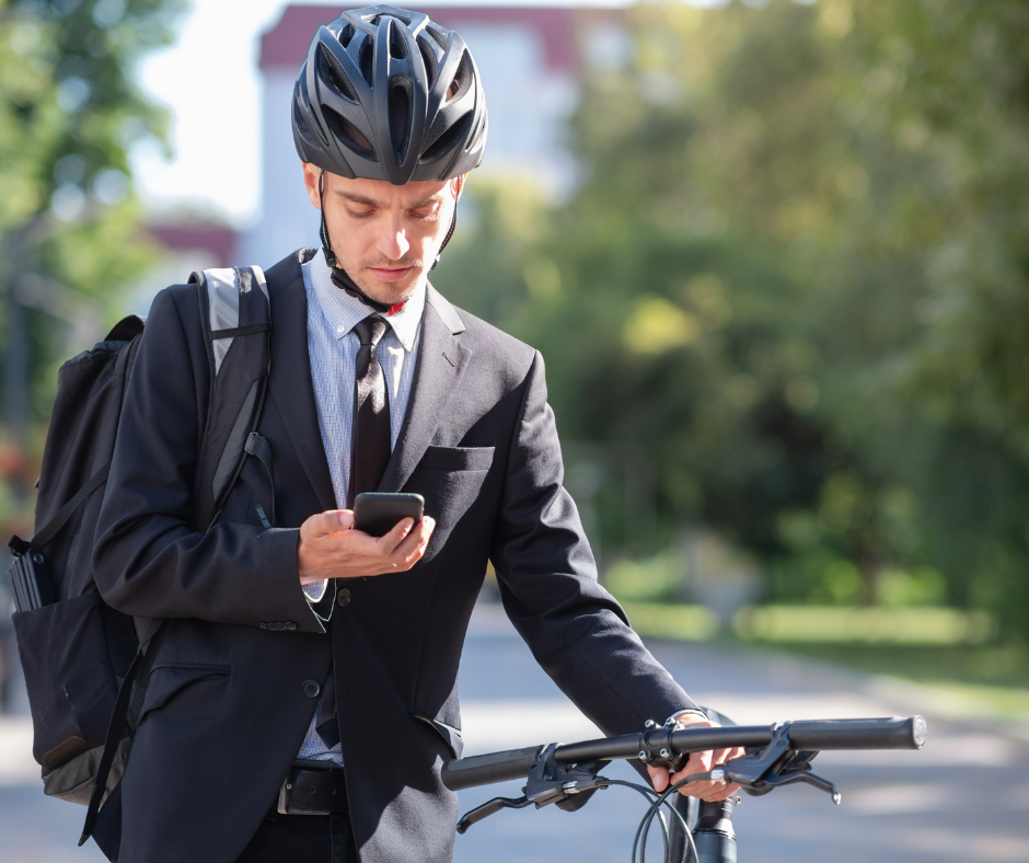 Un homme en costume et casque de vélo consulte son téléphone tout en tenant son vélo, en extérieur.