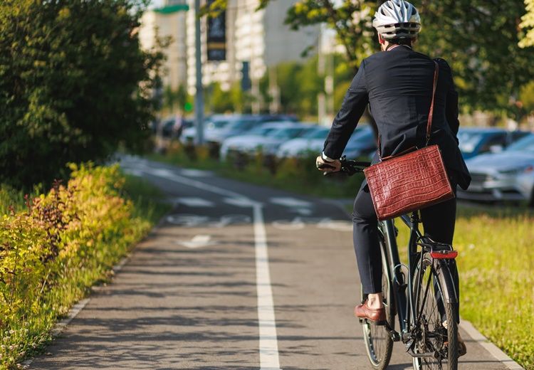 Homme en costume, casque et sac en bandoulière, roulant à vélo sur une piste cyclable bordée de verdure.