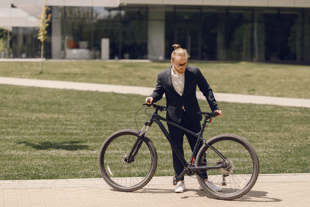 Un homme en costume élégant, portant des lunettes de soleil, marche à côté de son vélo tout-terrain devant un bâtiment moderne.