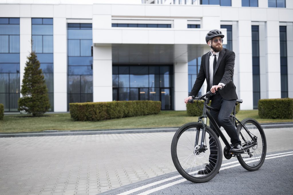 Un homme en costume et casque de vélo arrive à son travail à vélo, devant un bâtiment moderne aux façades vitrées.