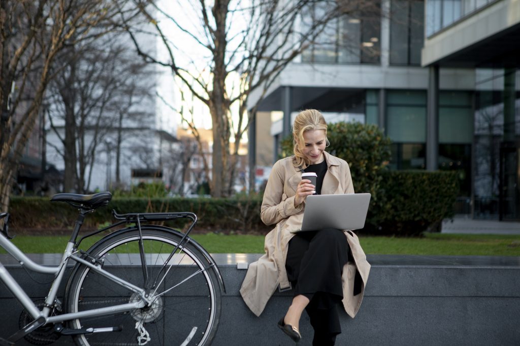 Une femme assise en extérieur près d'un vélo travaille sur son ordinateur portable en tenant un café dans un cadre urbain.