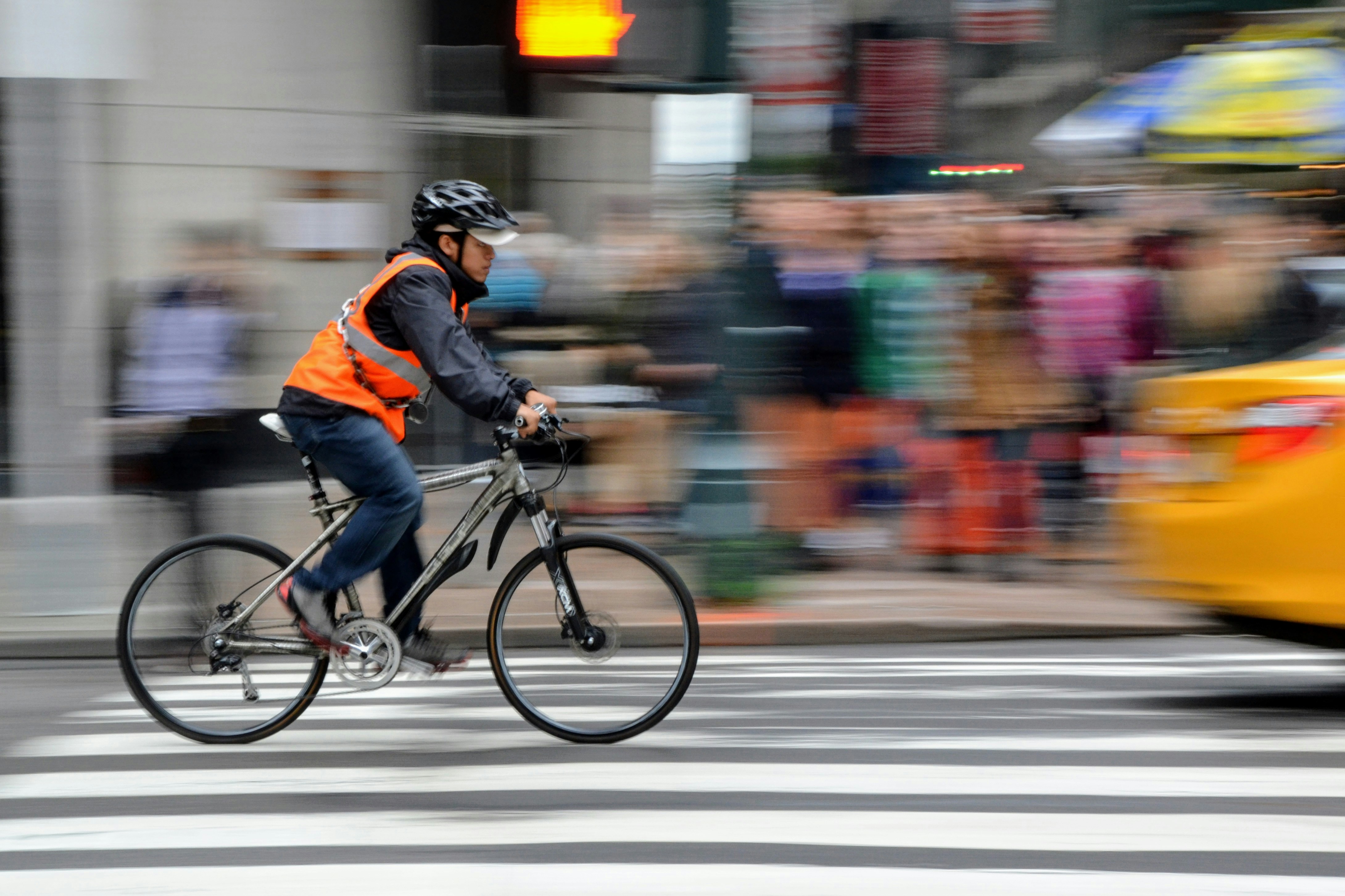 Un cycliste qui pédale en ville sur une route, avec un casque, un gilet orange, sur son vélo